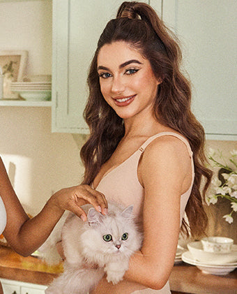 Woman wearing beige camisole holding a fluffy white cat in a kitchen setting.