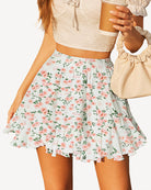 Model in a floral pleated mini skirt and sheer blouse, holding a green handbag.